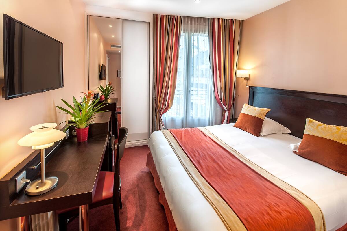 A hotel room with a bed, a desk with a lamp, a potted plant, and a TV on the wall.
