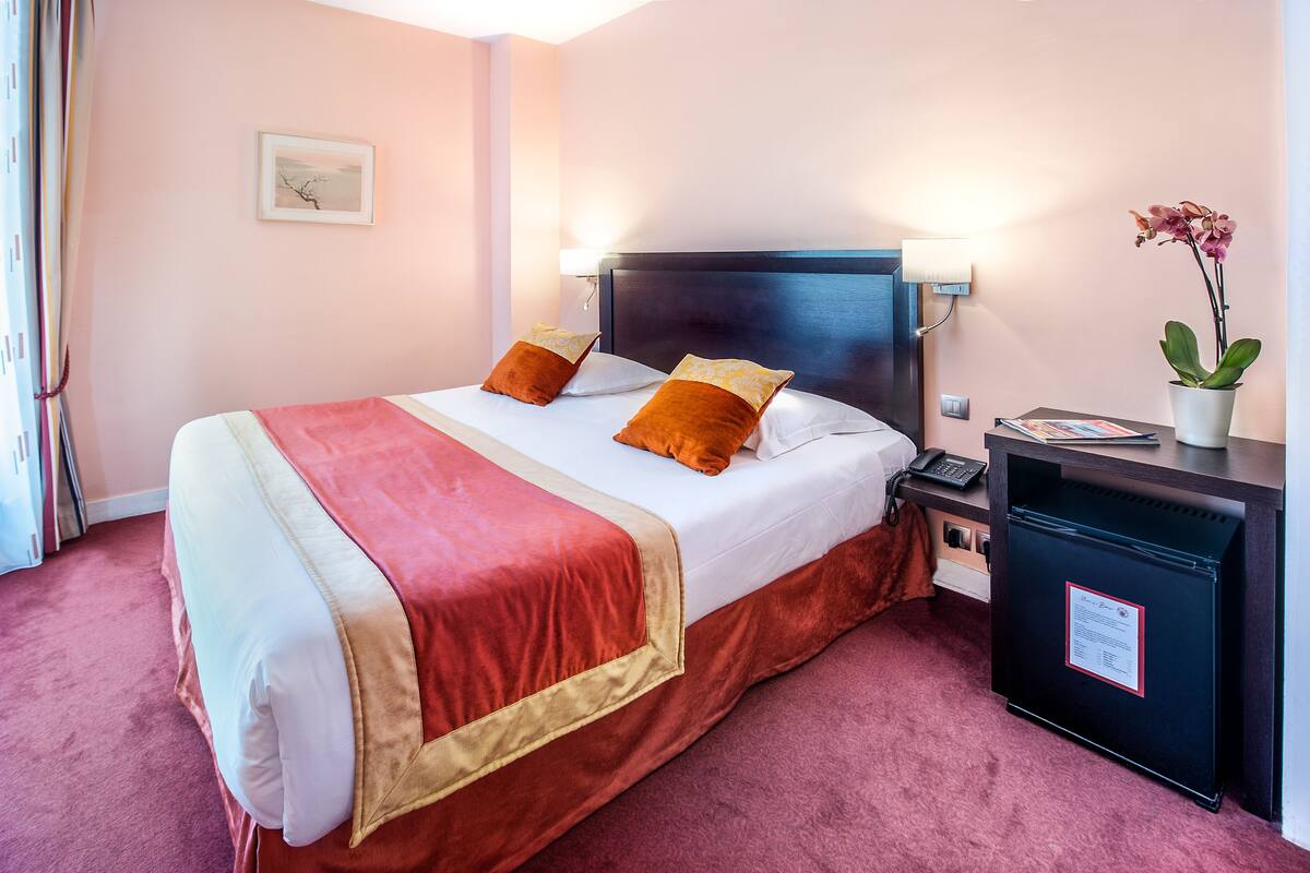 A hotel room with a bed, pillows, a bedside table with a lamp, a small refrigerator, and a potted plant.