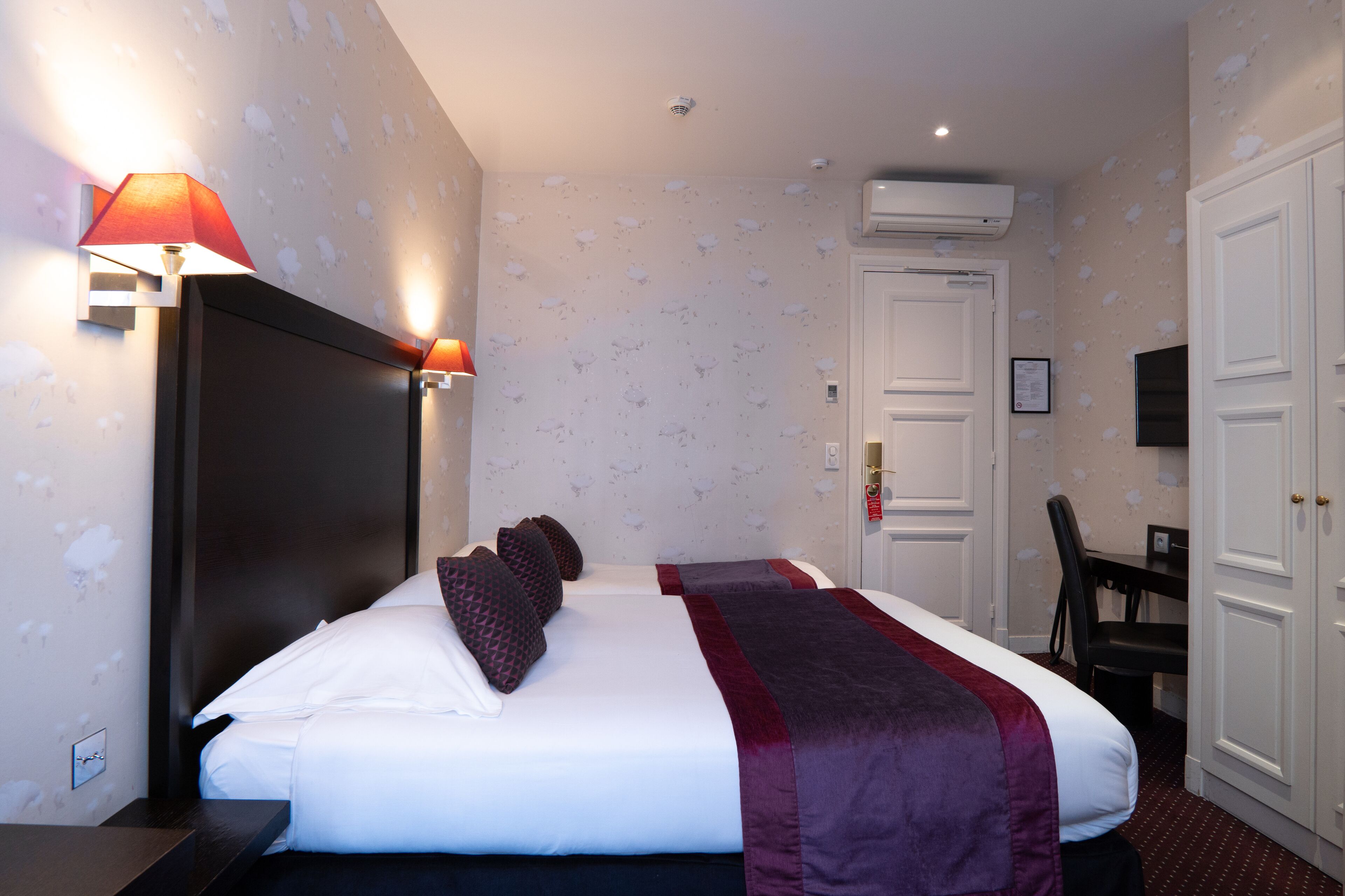 Une chambre d’hôtel avec un grand lit, un bureau et une chaise. On y trouve une lampe fixée au mur et un appareil de climatisation.