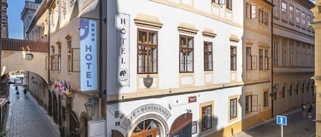 Un hotel con cafetería y restaurante en la planta baja, frente a una calle empedrada y con mesas al aire libre.