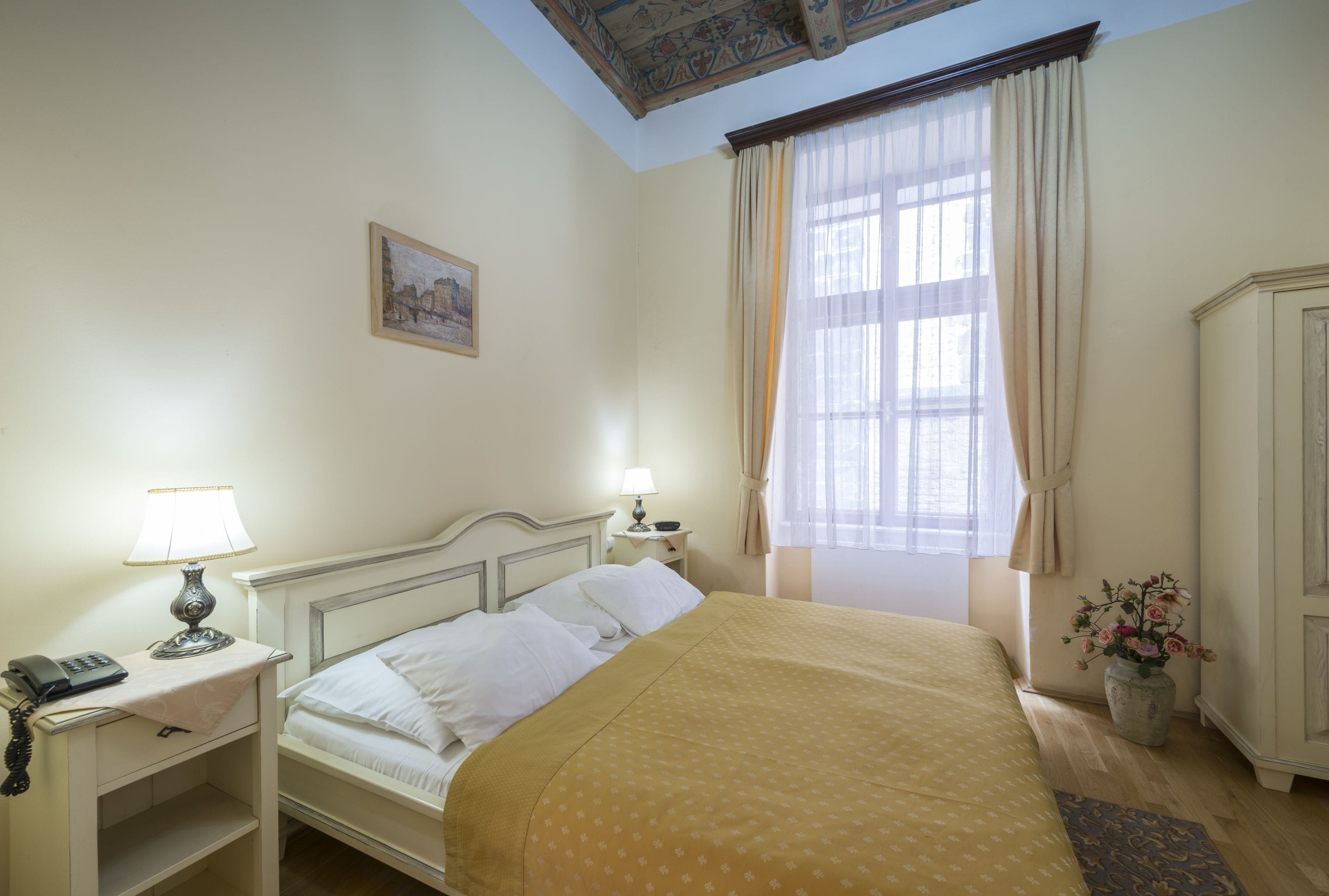 Habitación de hotel ordenada con cama, lámparas de noche, teléfono, una mesita y un jarrón con flores.