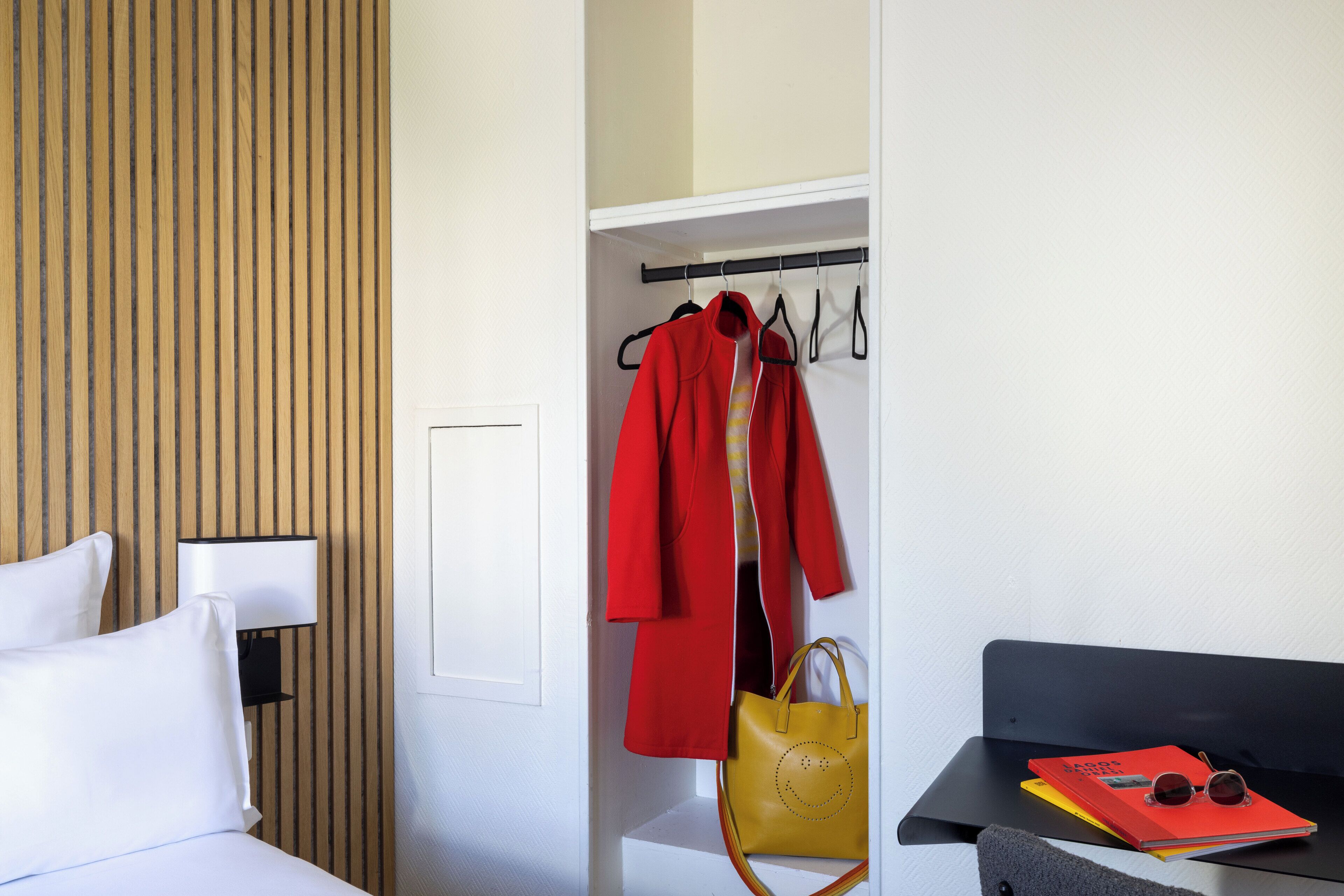 Habitación de hotel con cama, mesita de noche con lámpara, armario con ropa y bolso, y una pequeña mesa con un libro y gafas de sol.