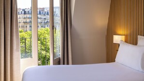 Una habitación de hotel con un ventanal grande, una cama, una mesita de noche y una lámpara fijada a la pared.