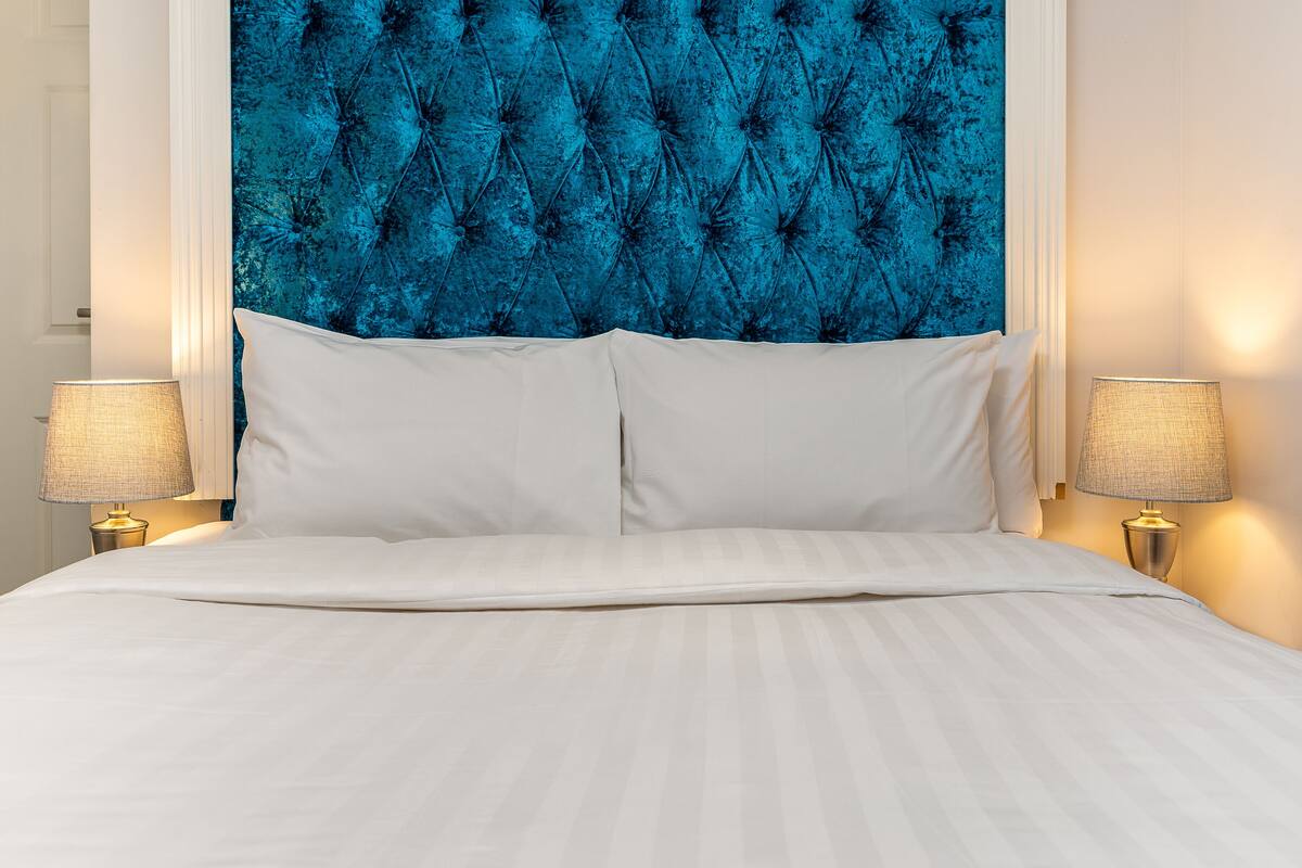 Ein Bett mit weißen Leinen, zwei Nachttischlampen und einem blauen, gepolsterten Kopfteil.