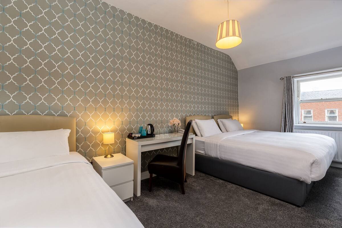 Ein Hotelzimmer mit zwei Betten, einem Schreibtisch, einem Sessel und einem Fenster mit Blick auf ein Gebäude.