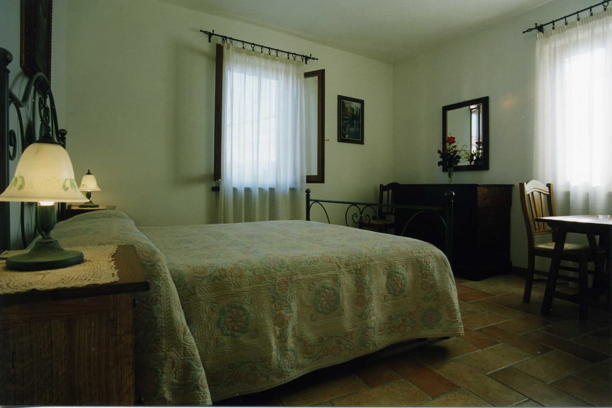 Ein Schlafzimmer mit Bett, Schreibtisch, Stuhl, Lampe, Kommode, Spiegel und einem Fenster mit Vorhängen.