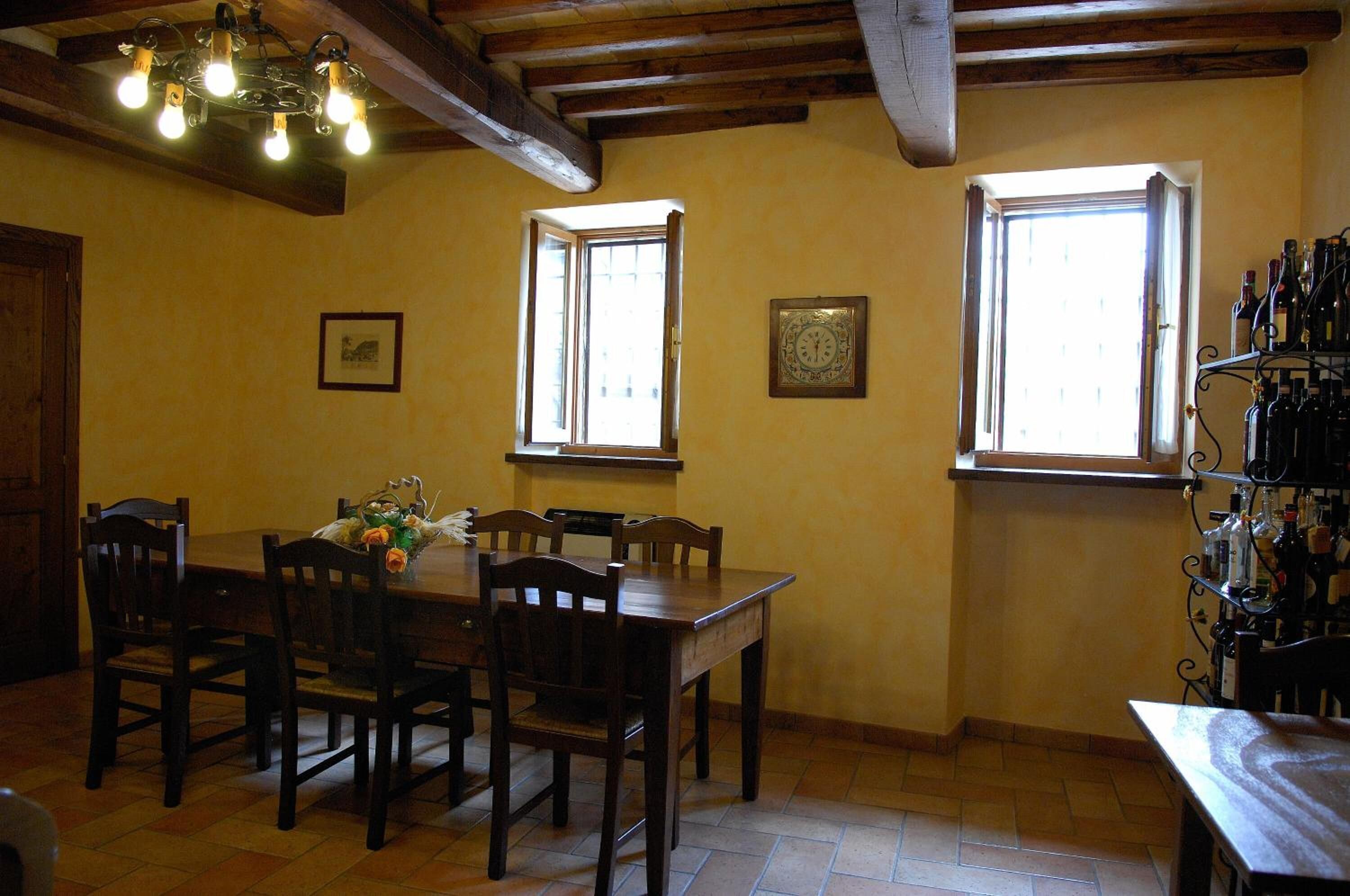 A dining area with a wooden table and chairs, two windows with curtains, a wall clock, and a wine rack with bottles.
