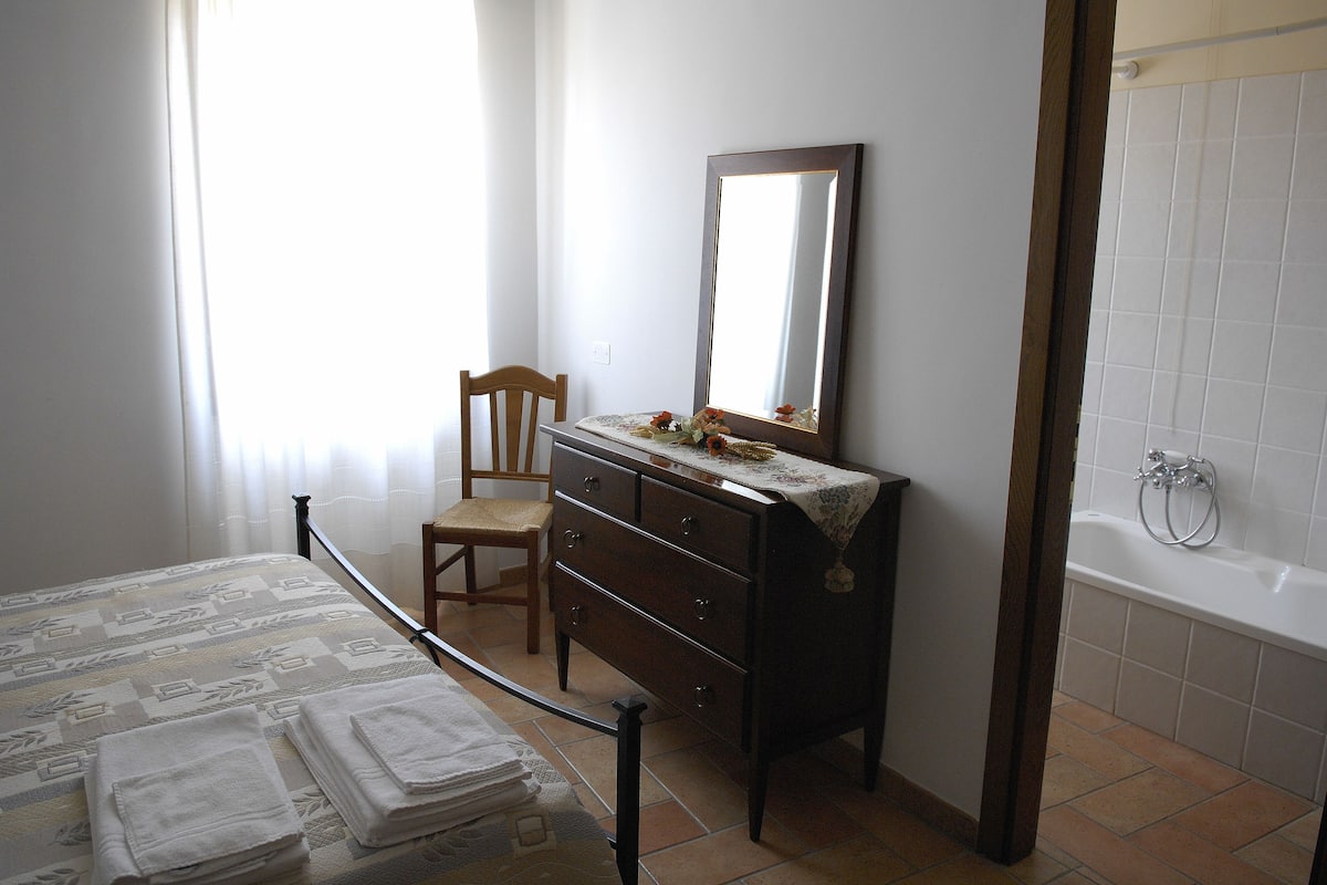 Ein Schlafzimmer mit Bett, Kommode, Stuhl und Spiegel, sowie ein Badezimmer mit Badewanne.