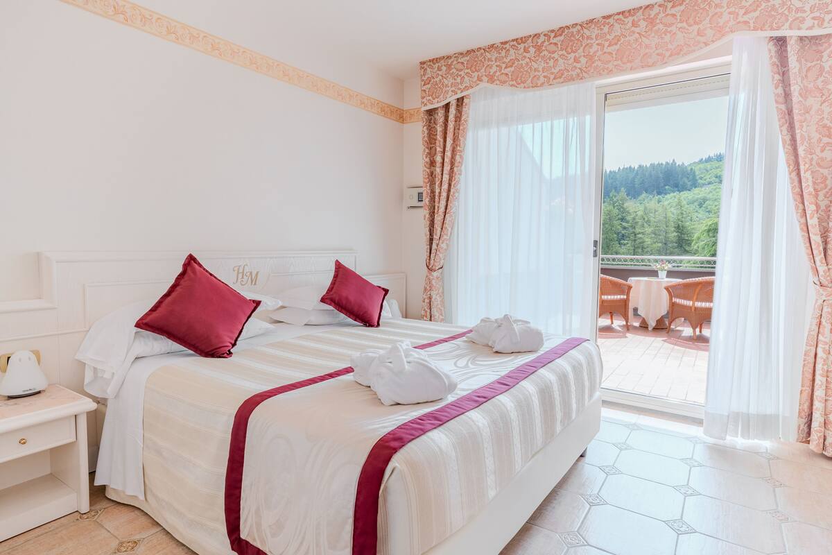 Una camera d'albergo con un letto grande, due cuscini rossi, una testiera bianca, un comodino con una lampada e un balcone con tavolo e sedie.