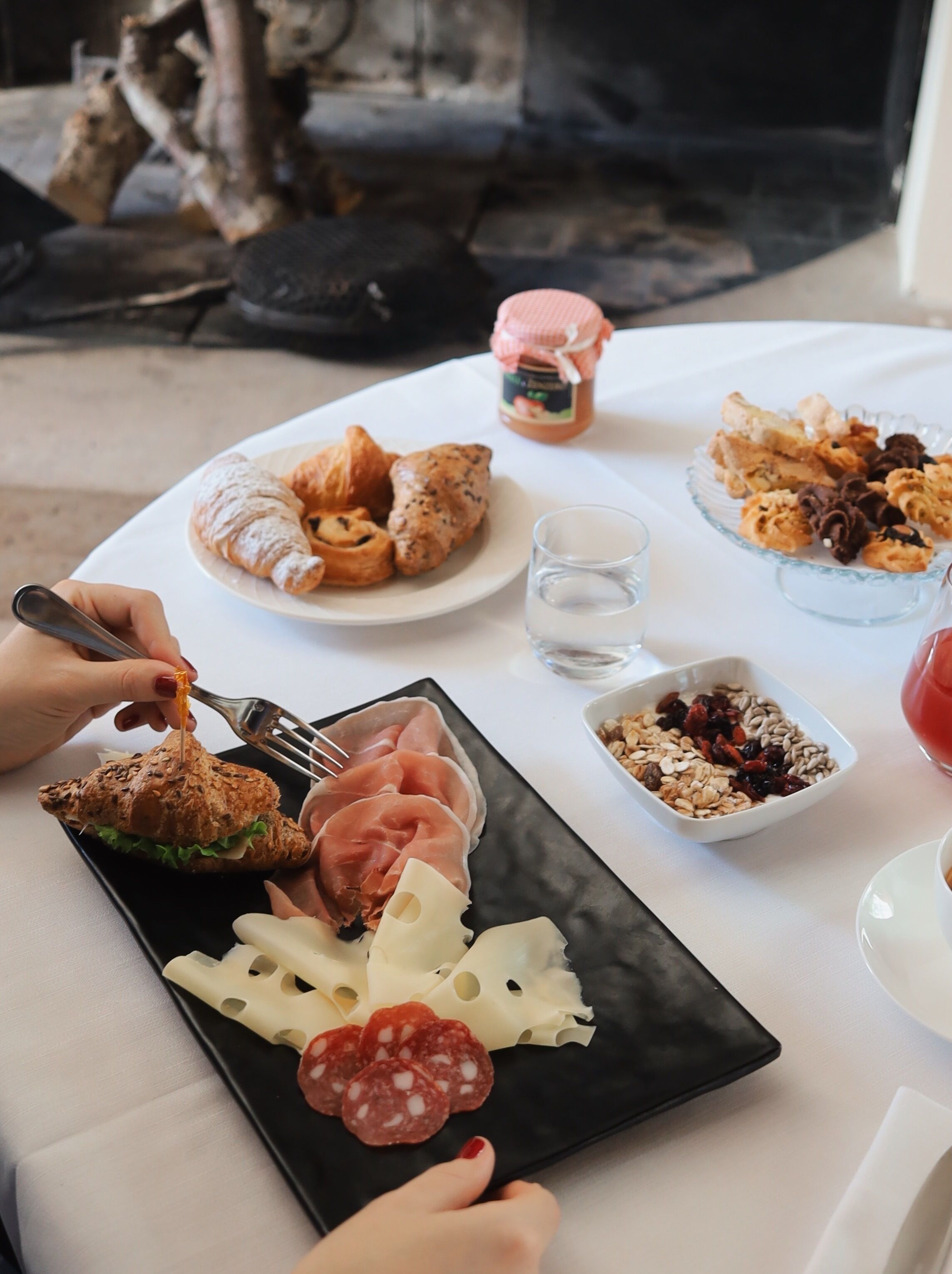 Une table est dressée avec divers aliments, notamment des croissants, un sandwich et une assiette de fromage et de salami. On peut également voir un pot de confiture et un bol de granola.