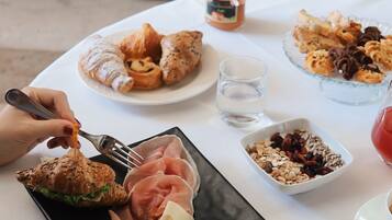 Ein Tisch mit verschiedenen Speisen, darunter Croissants, ein Sandwich und ein Teller mit Käse und Salami. Eine Marmeladenglas und eine Schüssel mit Granola sind ebenfalls zu sehen.