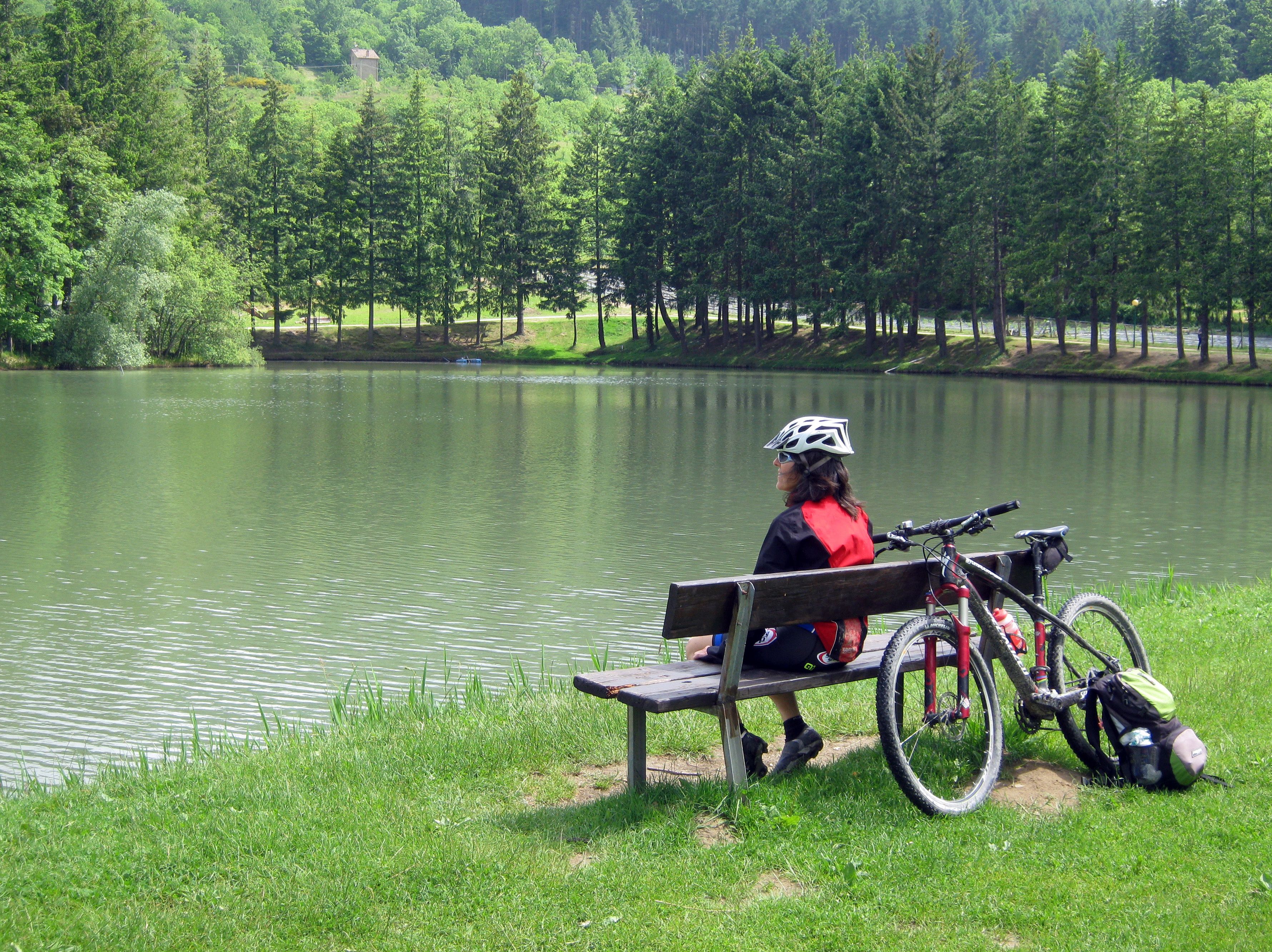 A person sitting on a bench by a lake with a bicycle and gear beside them.