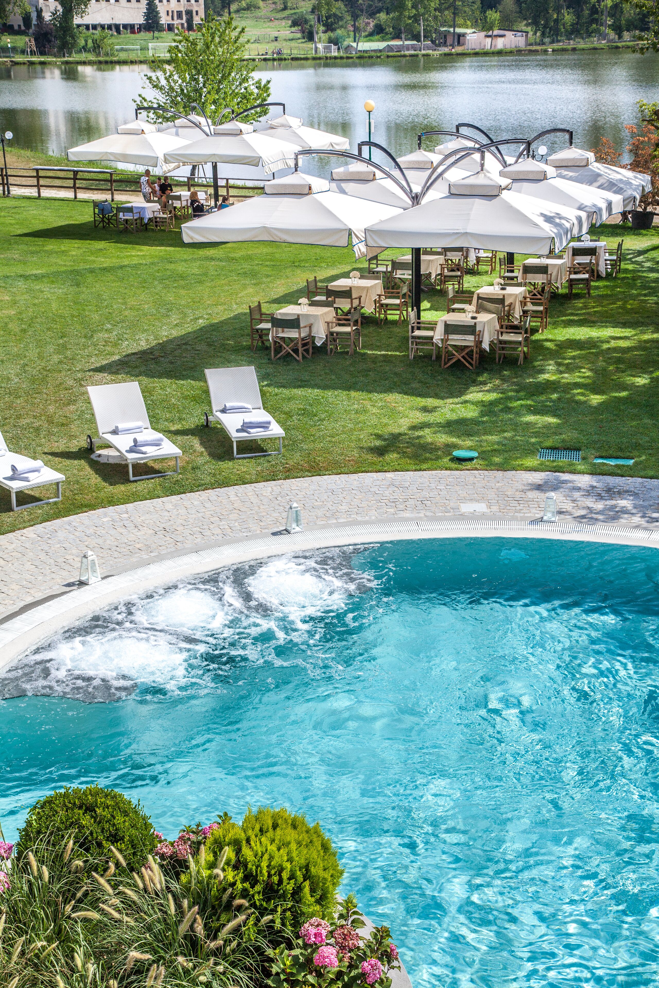 Un área de piscina con sillones de descanso y sombrillas, un jacuzzi y vistas a un lago.
