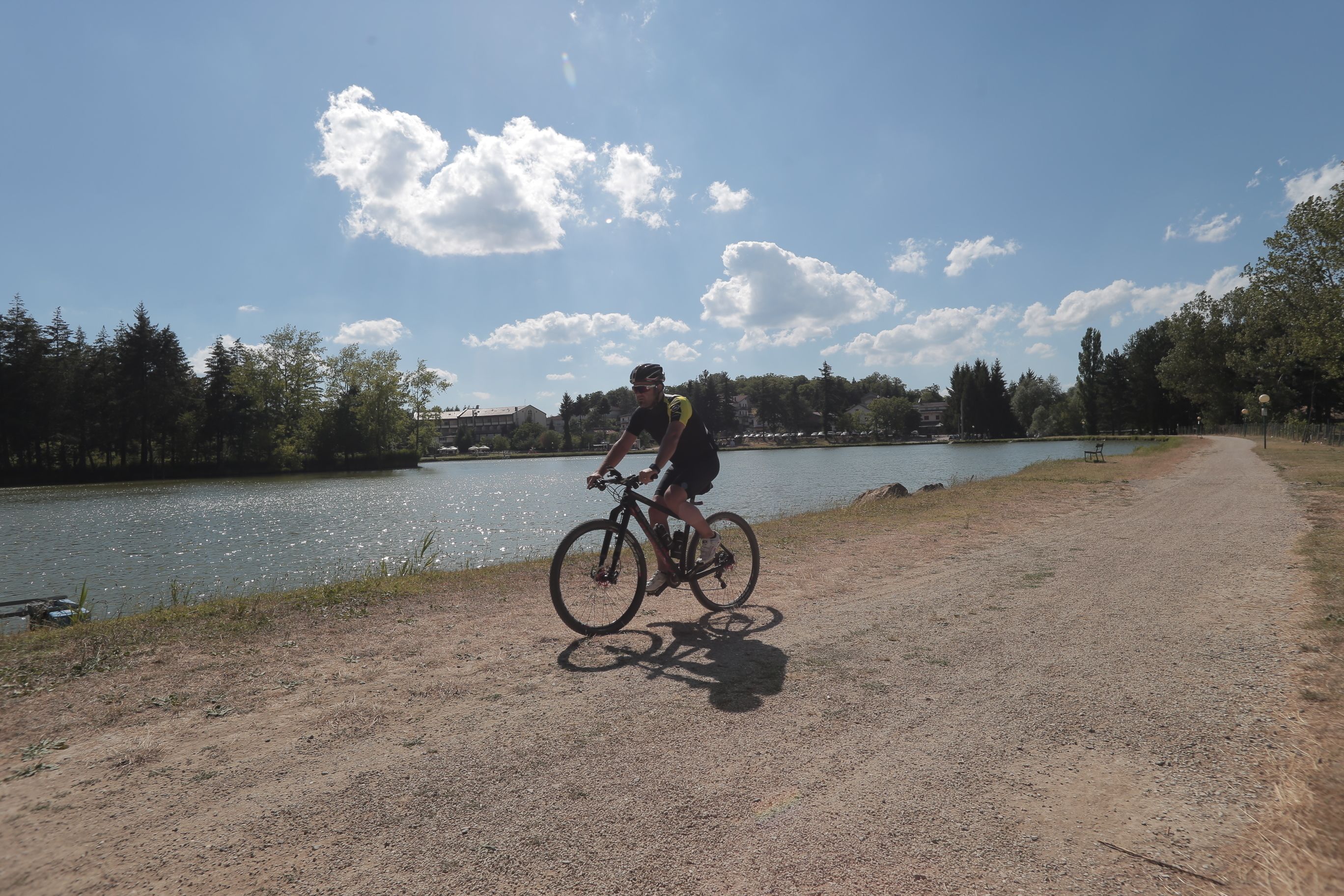 Eine Person fährt Fahrrad auf einem unbefestigten Weg neben einem Gewässer, im Hintergrund Bäume und Gebäude.