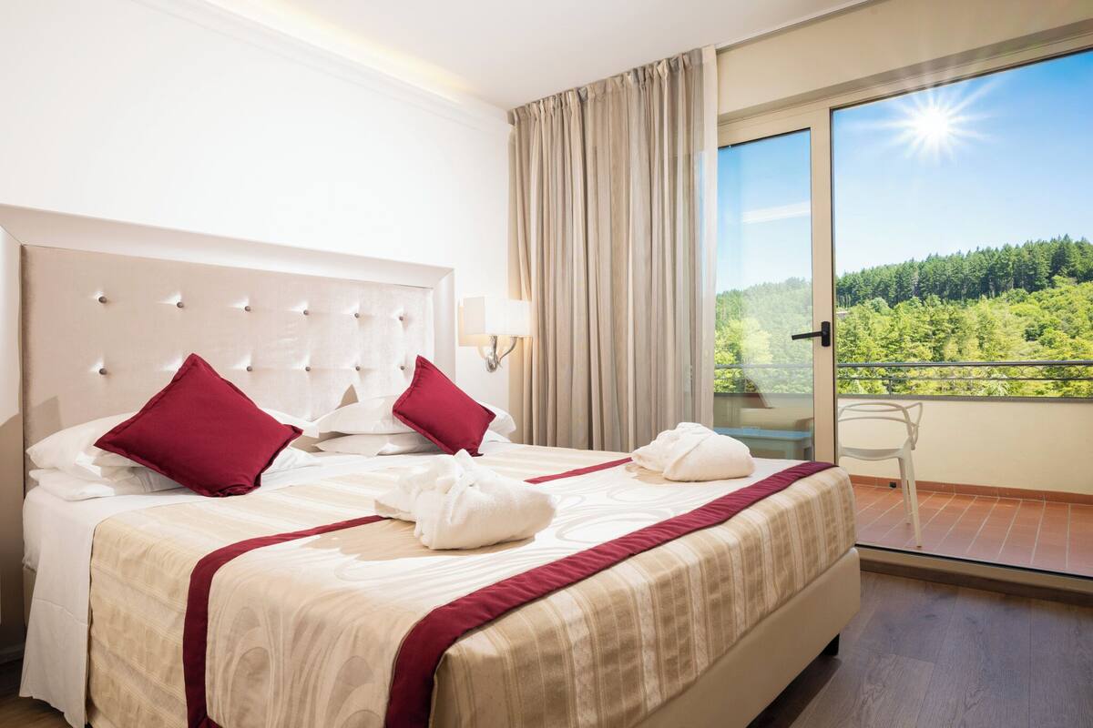 Camera d'albergo con un letto grande, due cuscini rossi, una testiera bianca, un balcone con vista sul verde e un cielo illuminato dal sole.