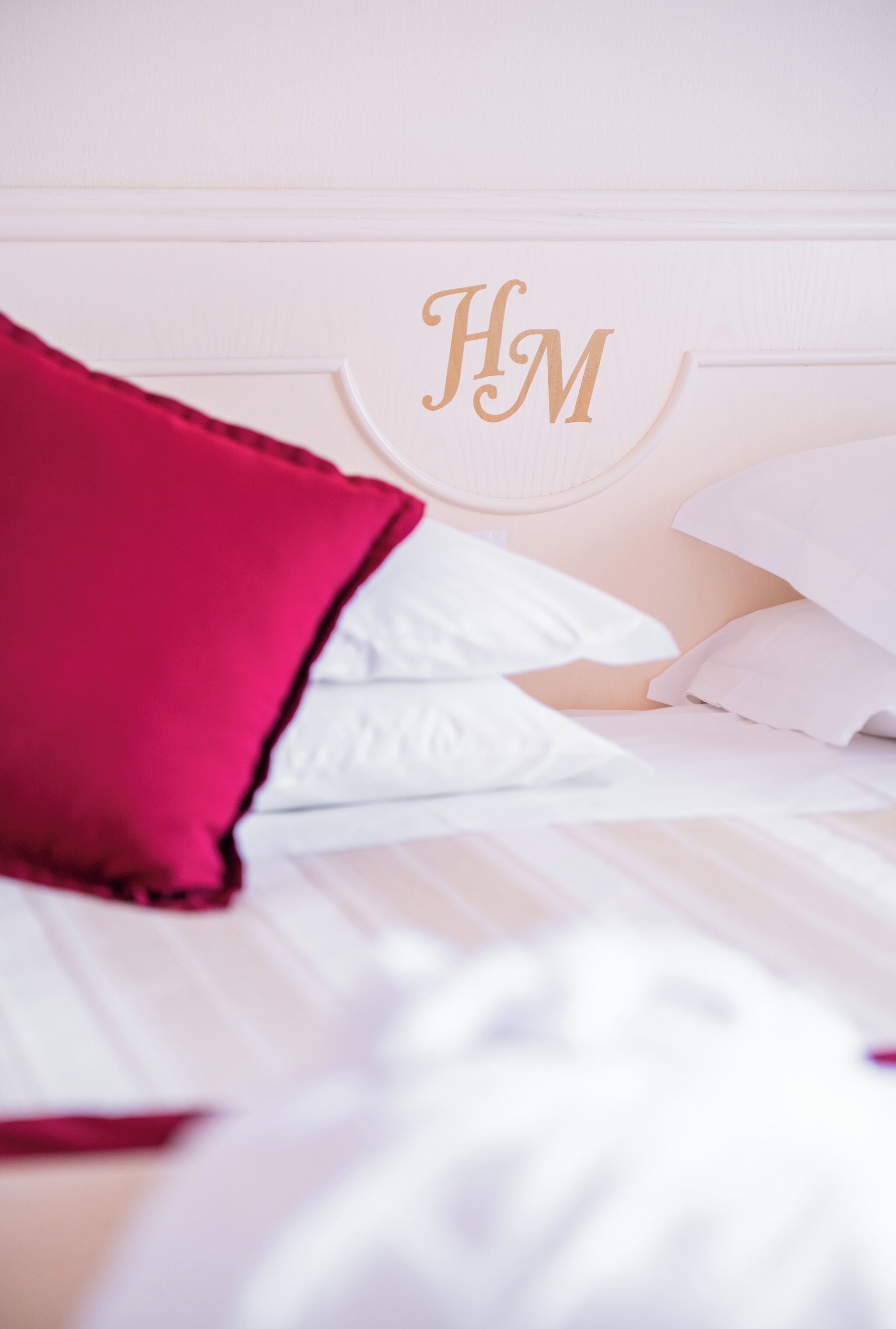Una cama con ropa de cama blanca y un almohadón rojo, un cabecero blanco con el monograma 'HM' en dorado y una pared blanca.