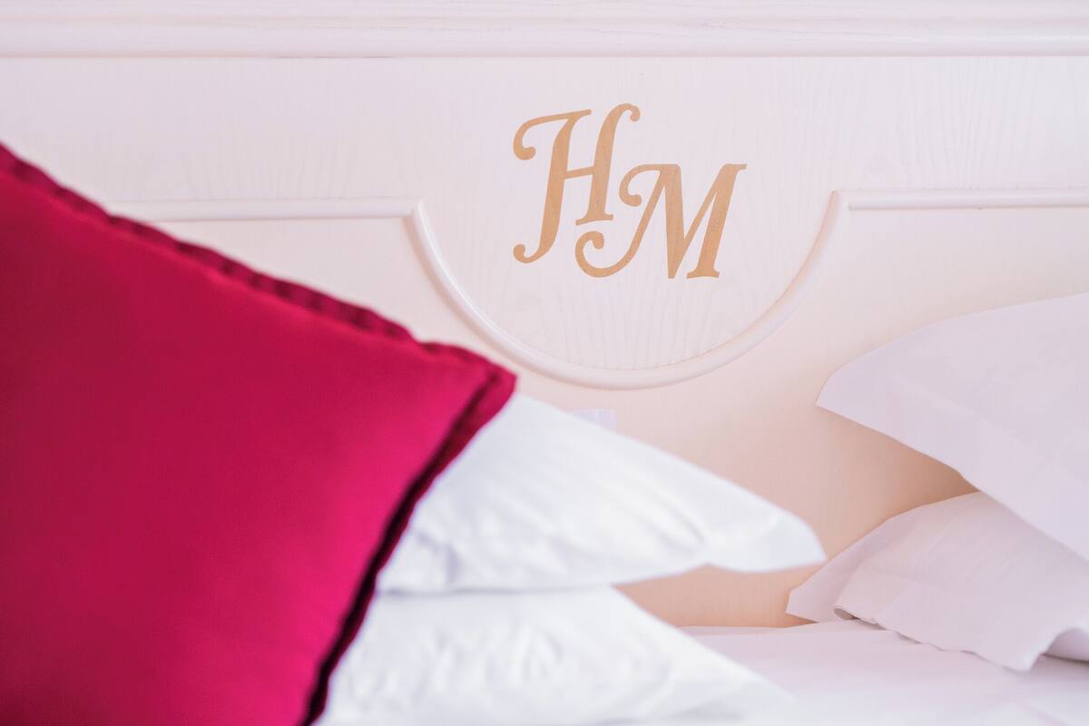 Un letto con biancheria bianca e un cuscino rosso, una testiera bianca con monogramma 'HM' dorato e una parete bianca.