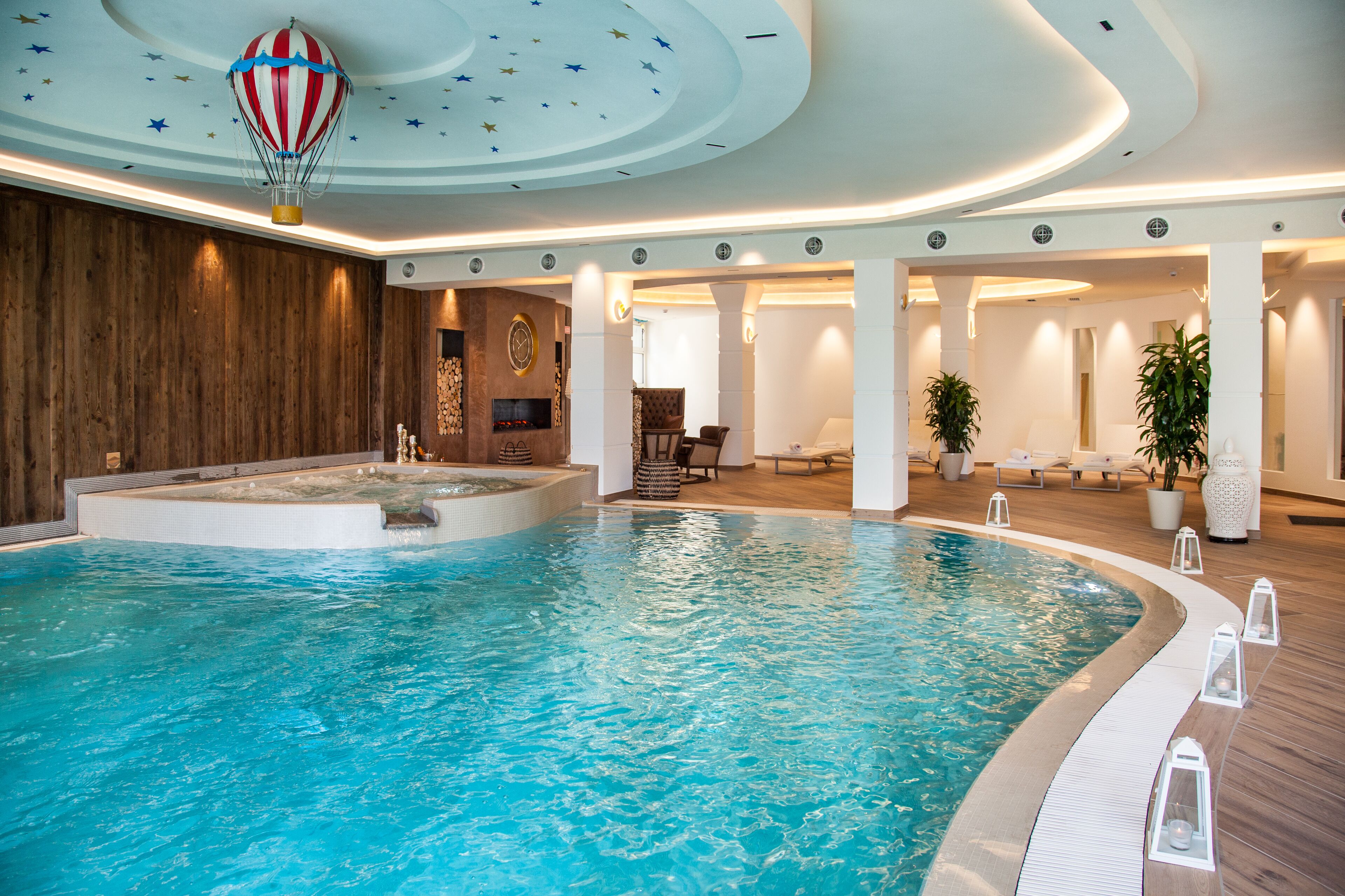 Uma grande piscina interior com decoração de balão de ar quente no teto, painéis de madeira e uma área de estar com cadeiras e um sofá.