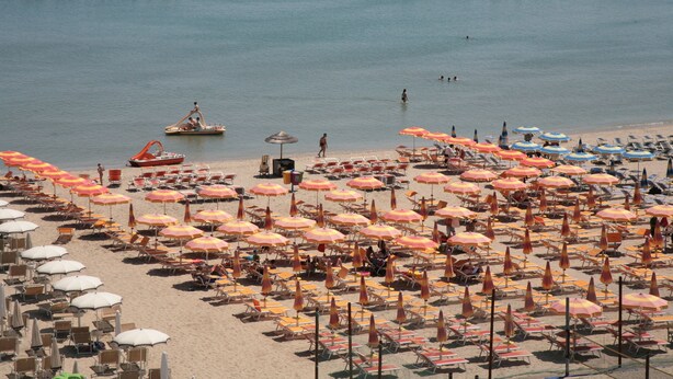 A beach with rows of sun loungers and umbrellas, a boat, and rocky outcrops in the water. - Residence San Paolo (Ravenna)