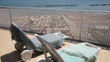 Ein Balkon mit zwei Liegestühlen und einem Tisch, der auf einen Strand mit Sonnenschirmen und Personen blickt.