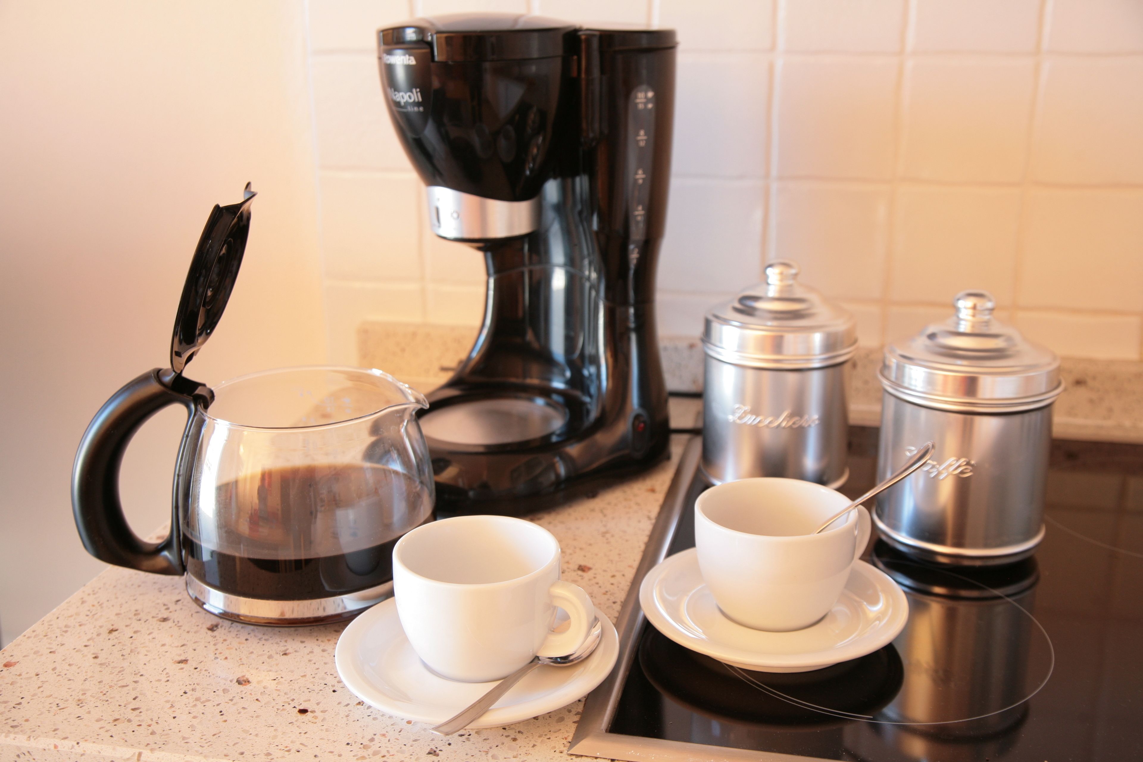 Una encimera de cocina con una cafetera, una jarra de vidrio, dos tazas blancas y dos contenedores de acero inoxidable.