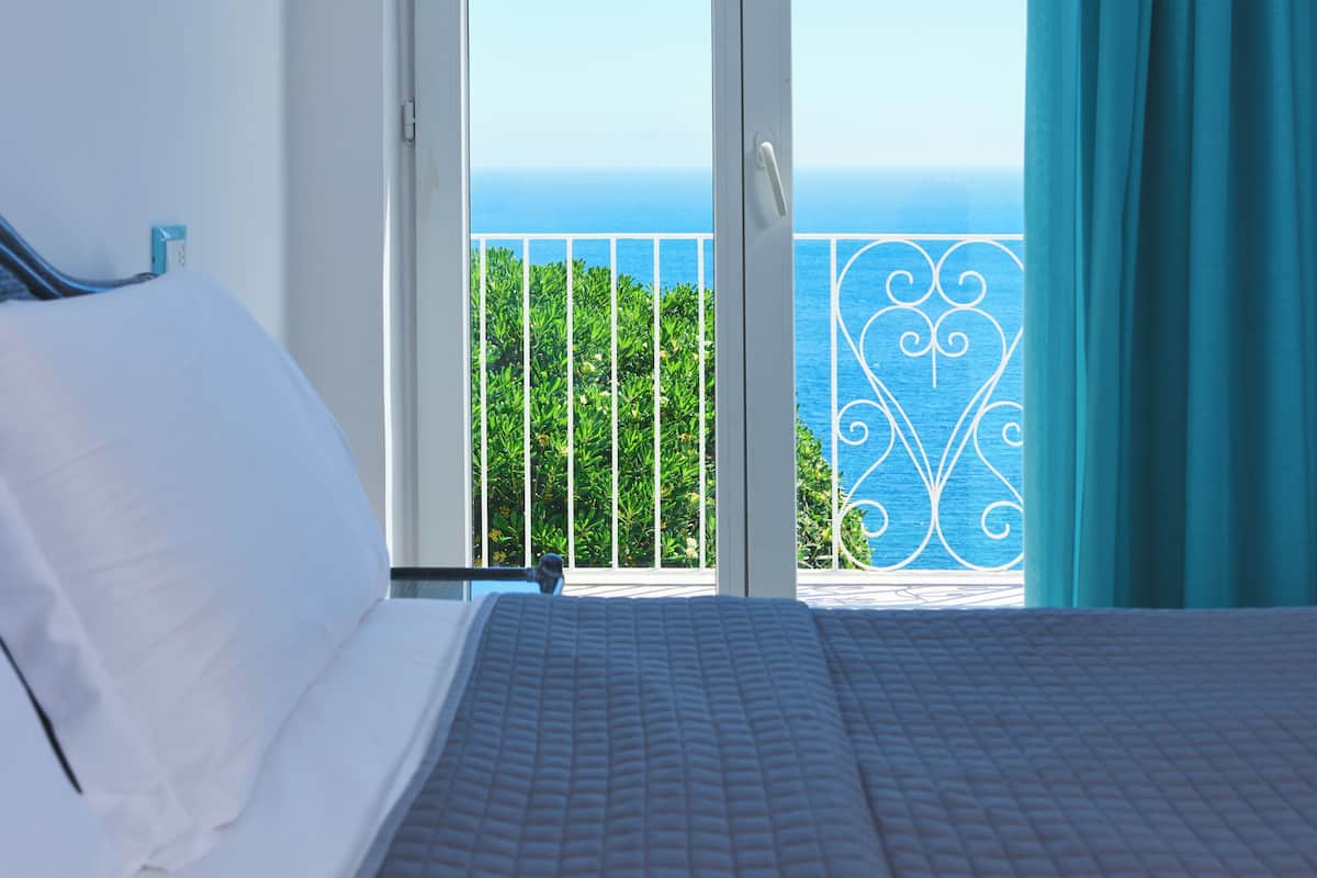 Una camera da letto con un letto, vista sul mare attraverso una porta vetrata e un balcone con ringhiera.