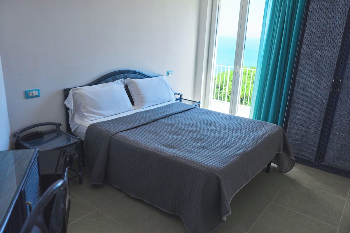 Camera d'albergo con un letto, una scrivania, una sedia, una finestra con tende e vista sul mare.