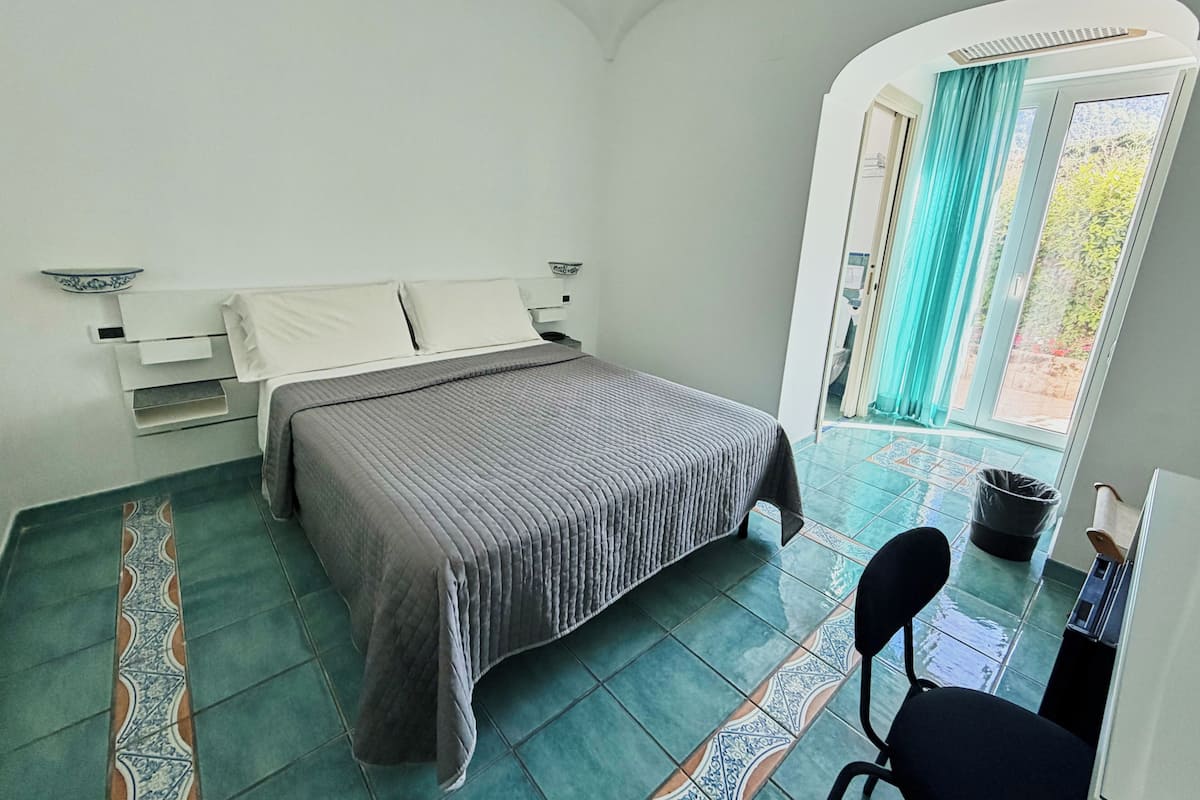 Una camera da letto con un letto, una sedia, una finestra con tende e un pavimento piastrellato.