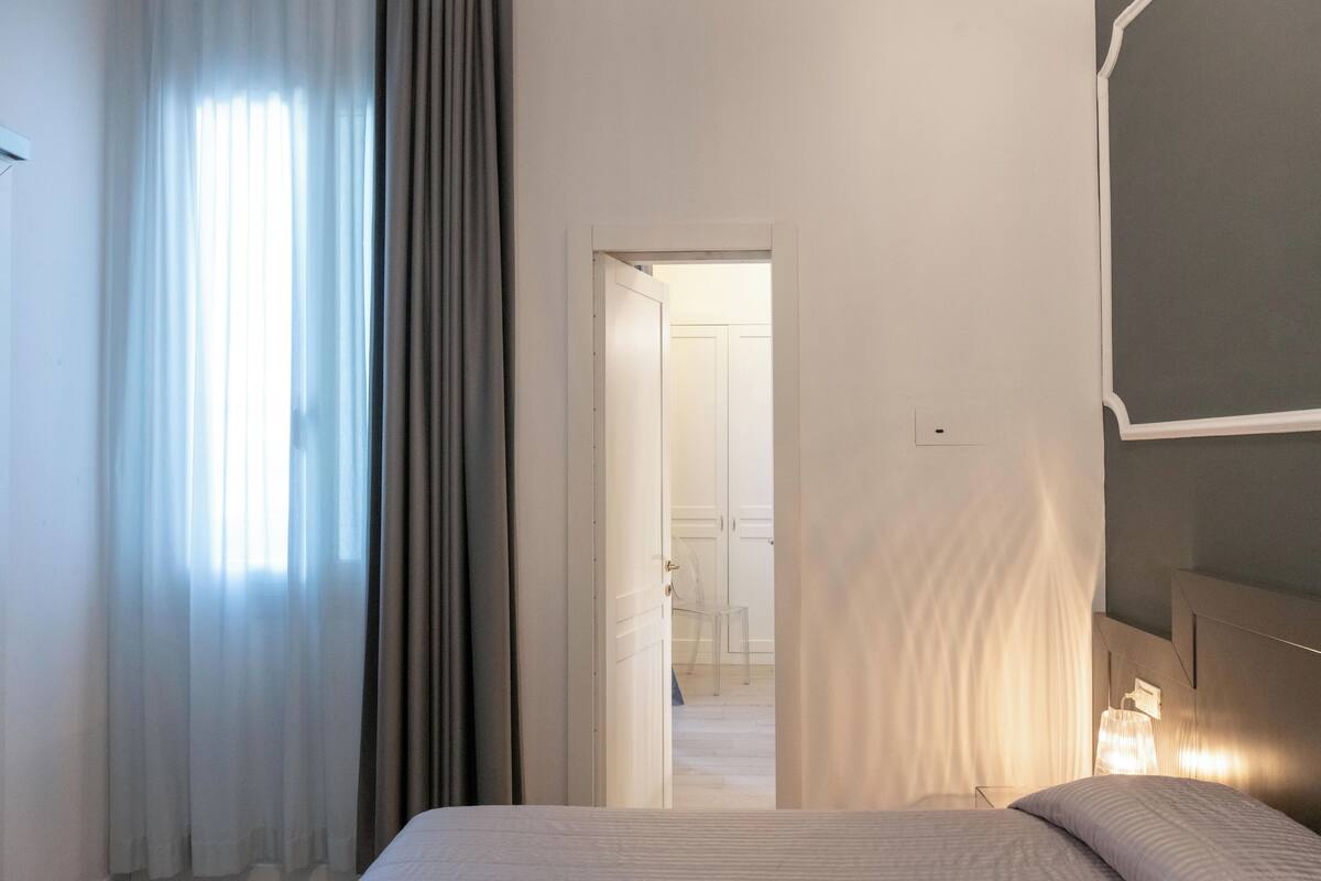 A hotel room with a bed, a nightstand, and a lamp. There is a door leading to another room.