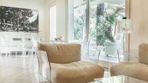 A modern living room with a beige sofa, clear chairs, a glass coffee table, and a large window with a view of greenery. - Perla Verde Hotel (Cervia)