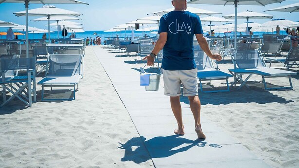A beachside area with rows of sun loungers and umbrellas, a man walking with a bucket, and a clear view of the ocean. - Perla Verde Hotel (Cervia)