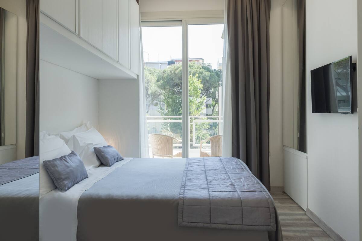 A modern hotel room with a large bed, a TV mounted on the wall, and a balcony with outdoor furniture.