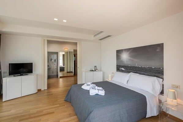 A hotel room with a bed, a television on a cabinet, a painting on the wall, and a view of the sea.