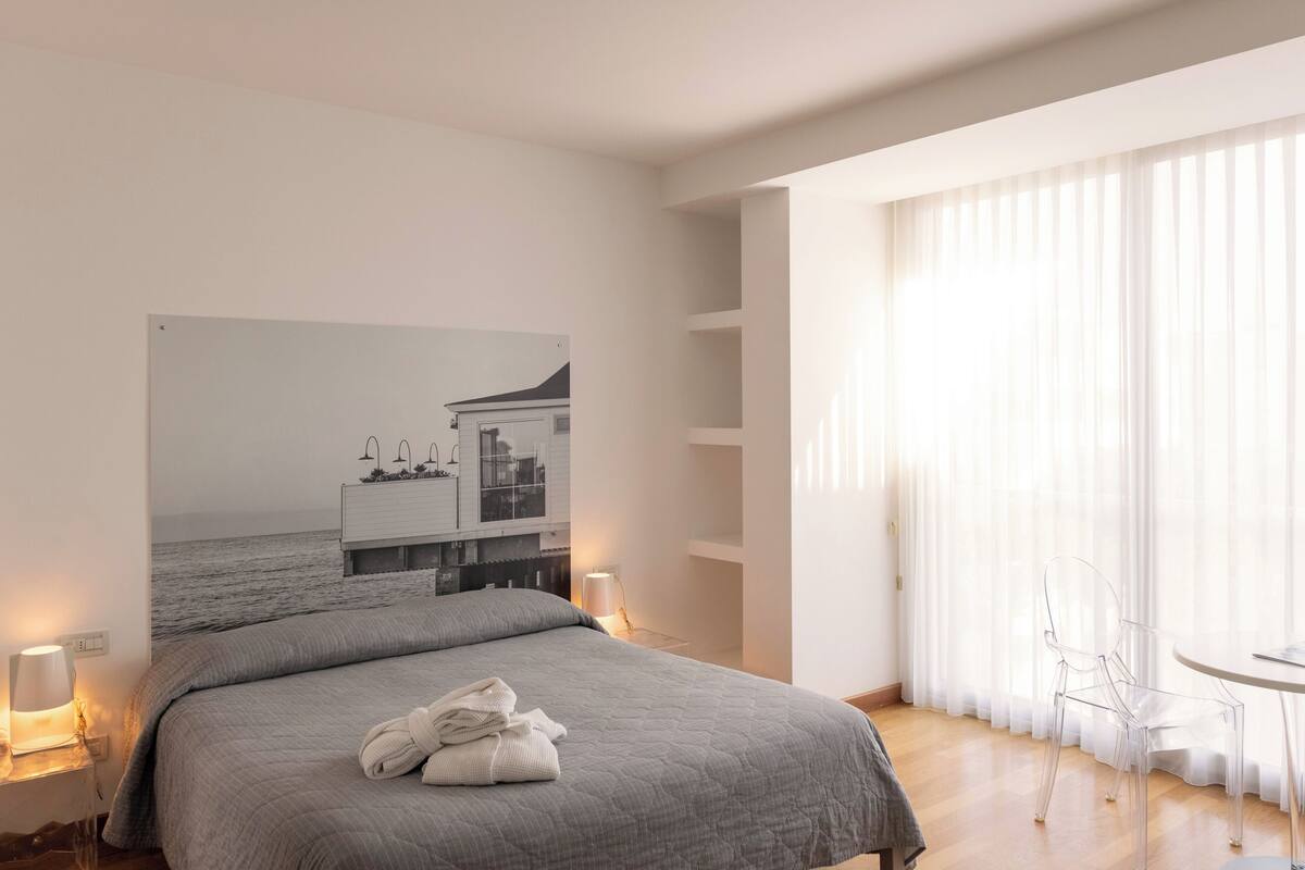 A modern hotel room with a large bed, a view of the sea, a dining area, and a bathroom.