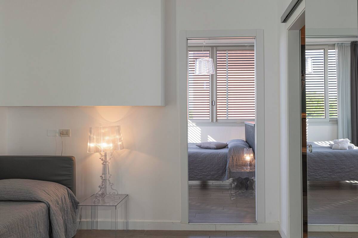 A bedroom with a bed, a bedside table with a lamp, and a view through a glass door to another room.