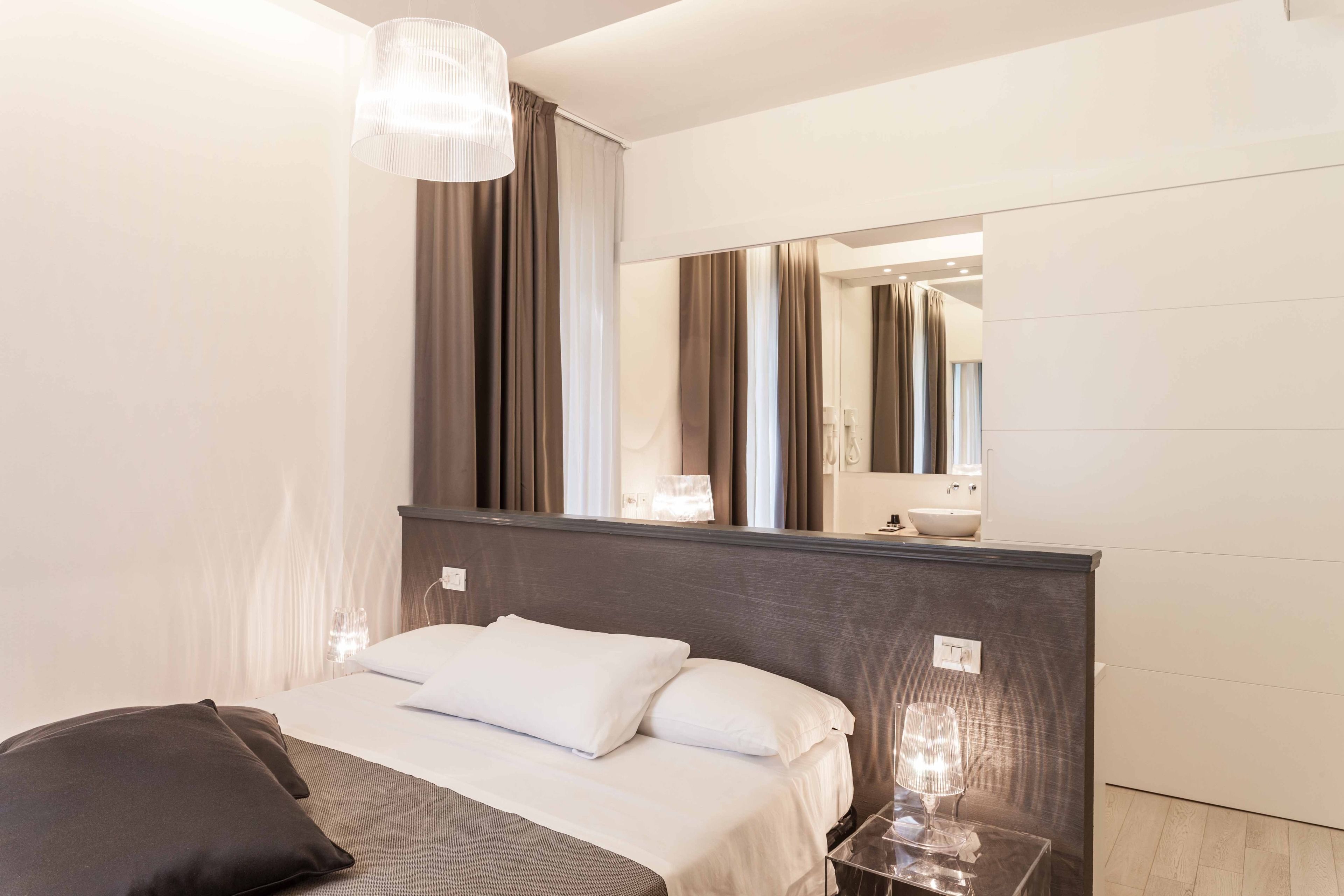A modern hotel room with a large bed, a bedside table with a lamp, a mirror, and a view of the bathroom.