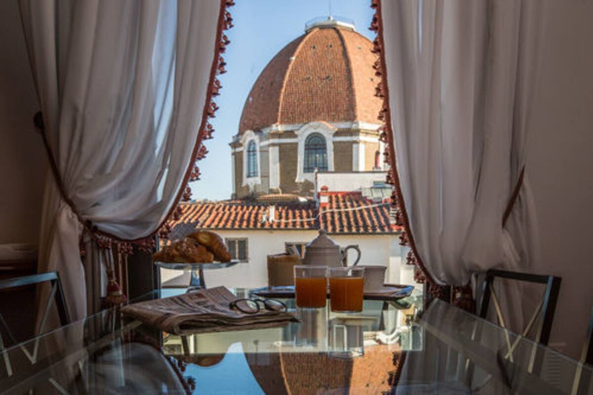 A dining table with a view of a building with a dome and tiled roof through sheer curtains.