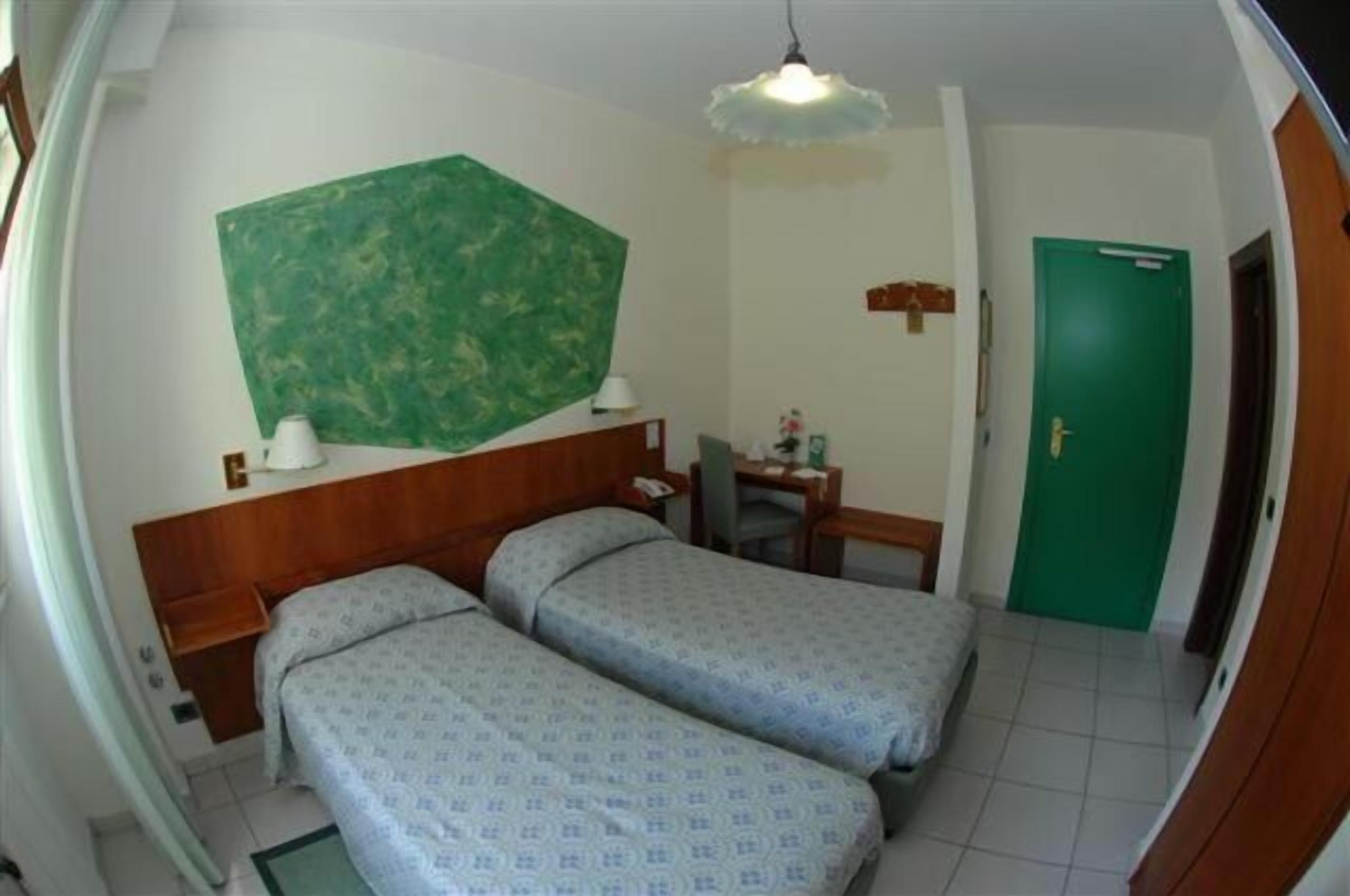 Une chambre avec deux lits, un bureau, une chaise, une porte verte et une lampe fixée au mur.