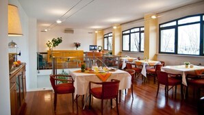Un comedor con varias mesas y sillas, una cocina al fondo y ventanas amplias con vistas a árboles desnudos.
