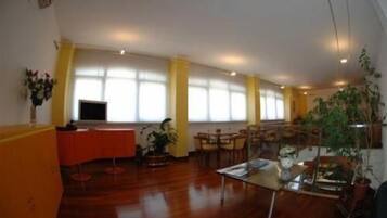 A spacious room with a wooden floor, a glass table, and multiple chairs. There are large windows with yellow curtains, a television, and several potted plants.