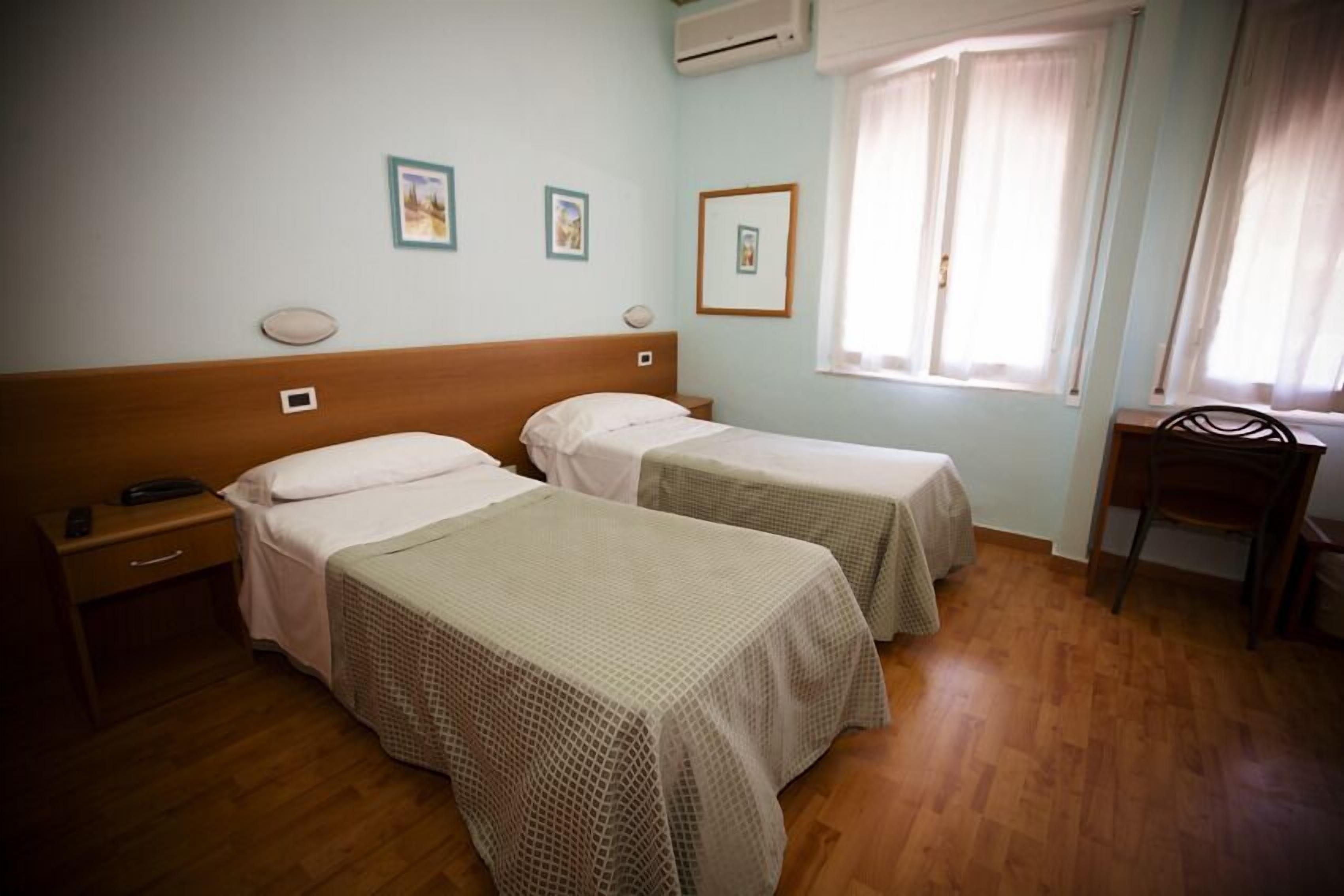 A room with two single beds, a wooden nightstand with a phone, a chair, and a window with curtains.
