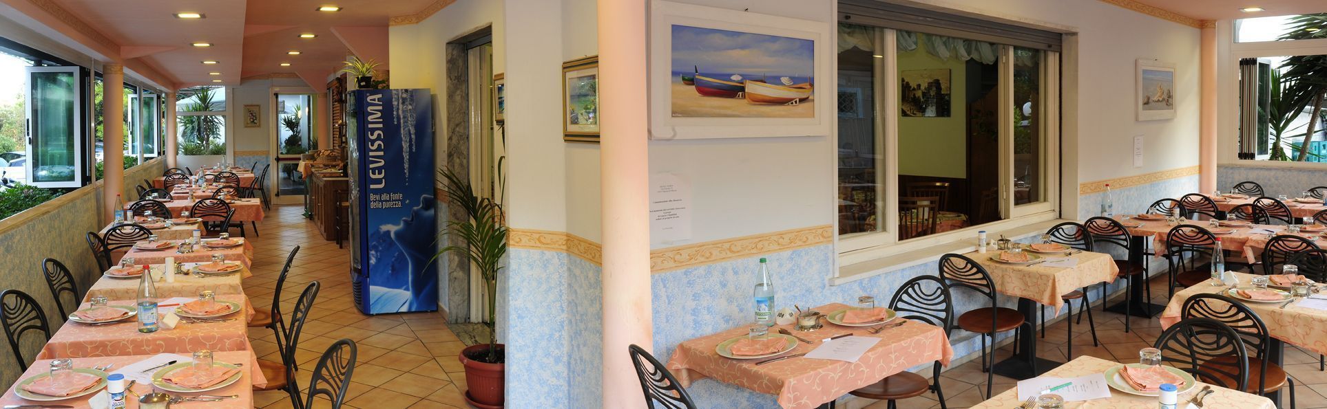 Un restaurant avec plusieurs tables dressées pour le service, une enseigne bleue portant l’inscription « LEVISIMA », et un tableau représentant des bateaux accroché au mur.