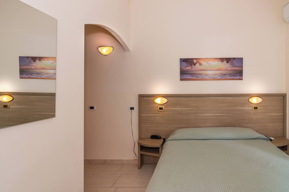 A hotel room with a bed, a nightstand, a wall-mounted lamp, and two paintings.