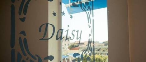 A room with a window view of outdoor tables and chairs, a wall with abstract cut-out designs, and the word "Daisy" prominently displayed.