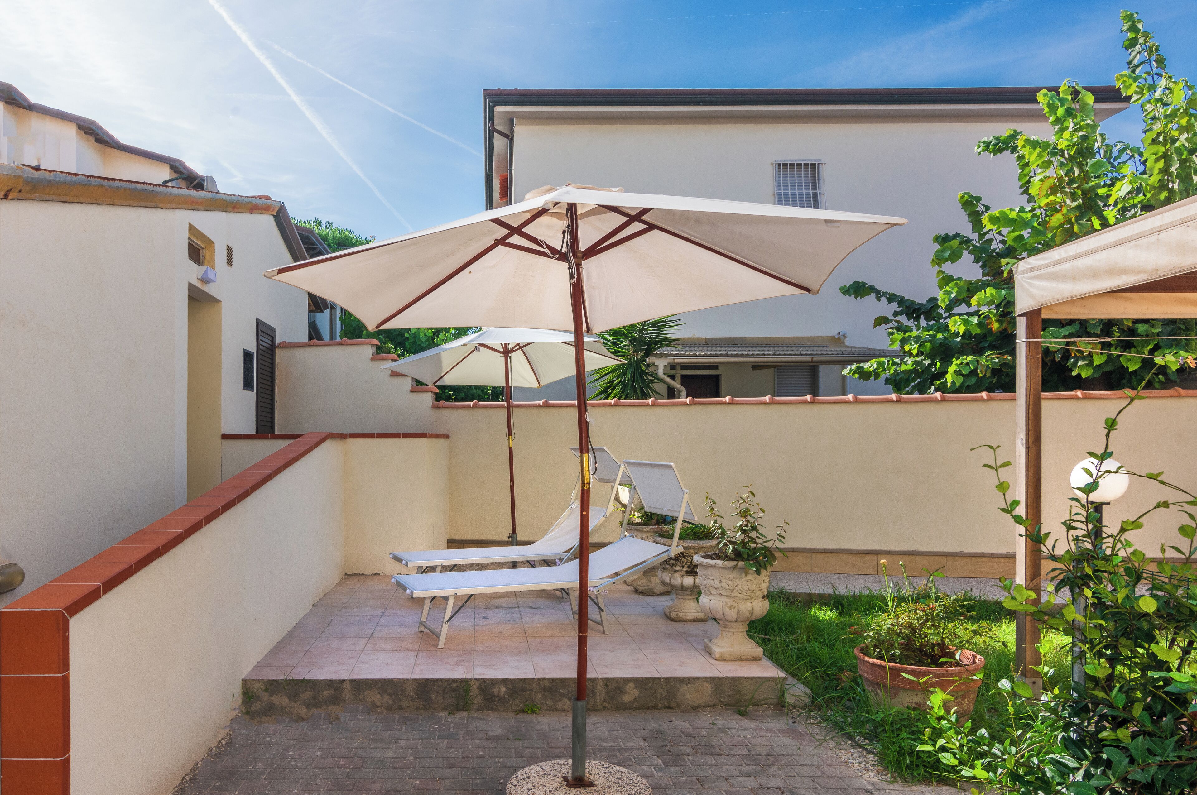 A patio with a sun lounger and a large umbrella, surrounded by potted plants and a building in the background.