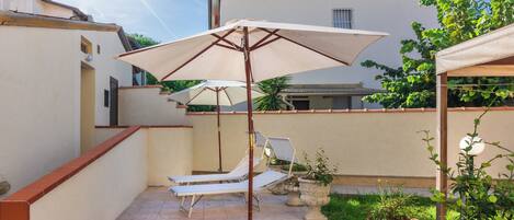 A patio with a sun lounger and a large umbrella, surrounded by potted plants and a building in the background.