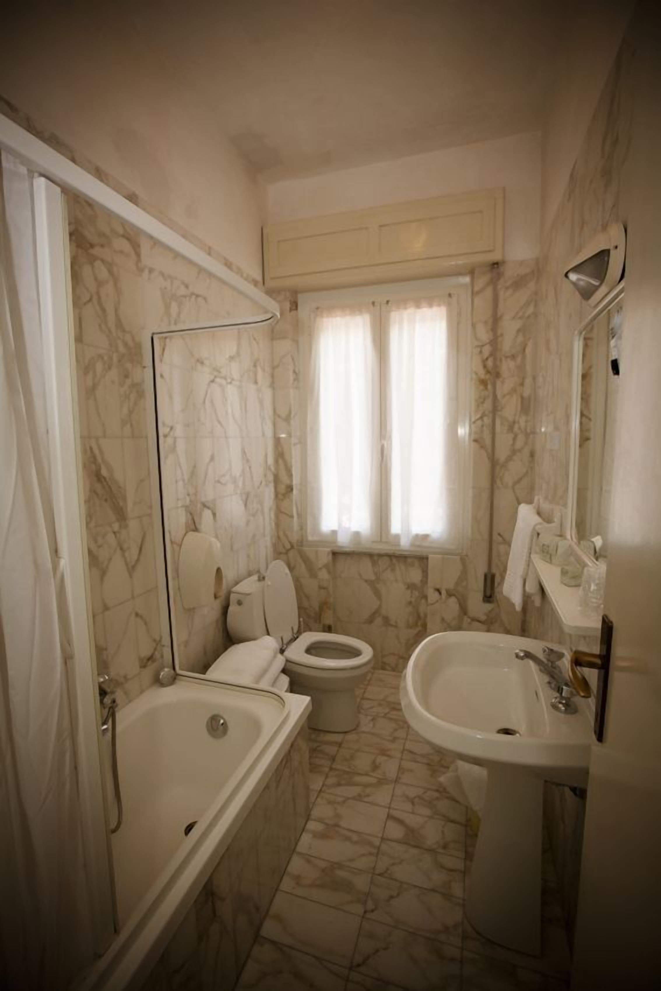 A bathroom with a bathtub, toilet, and sink, marble walls, and a window with curtains.