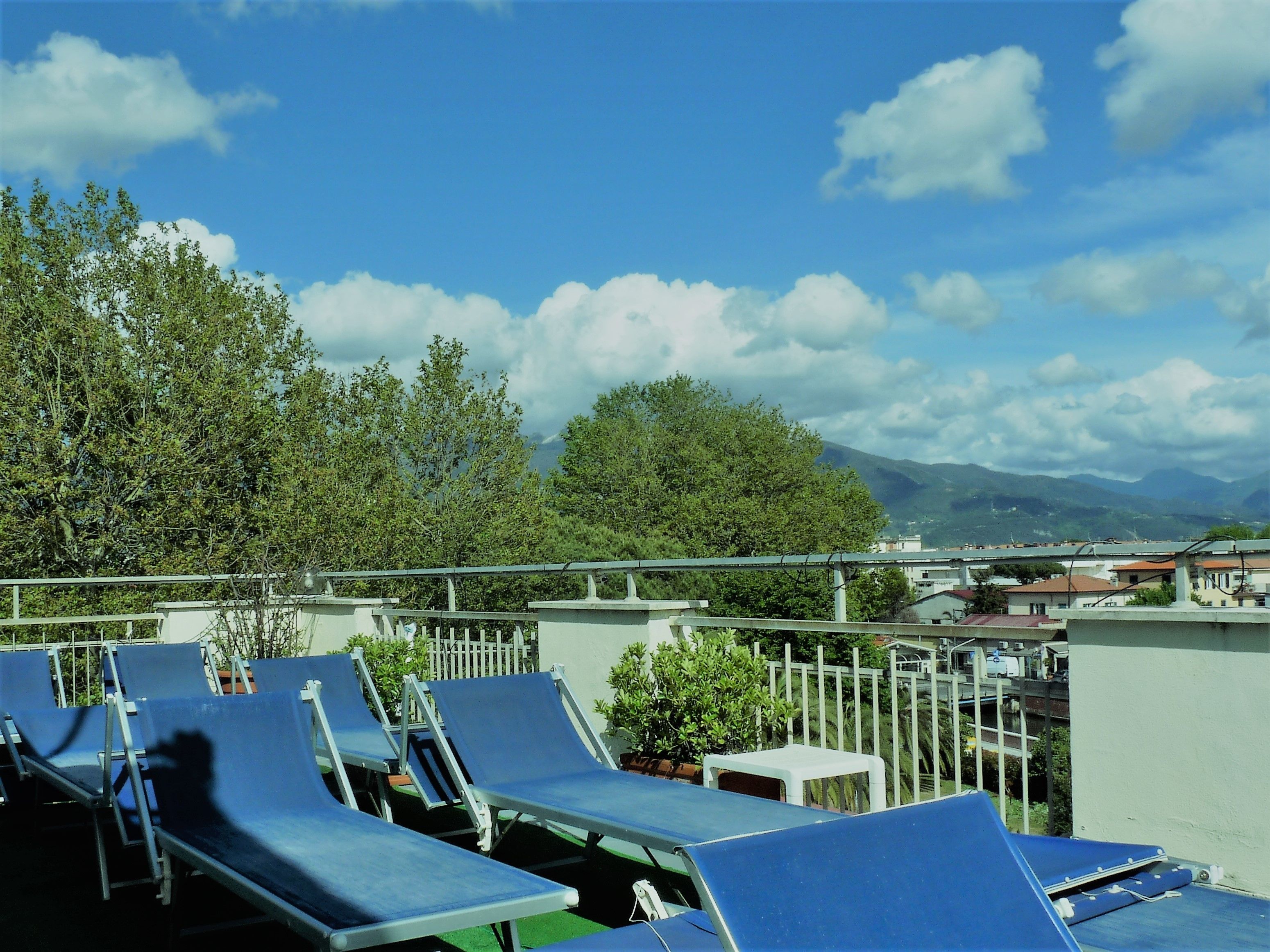A rooftop terrace with blue lounge chairs, a white fence, and a view of mountains and trees.