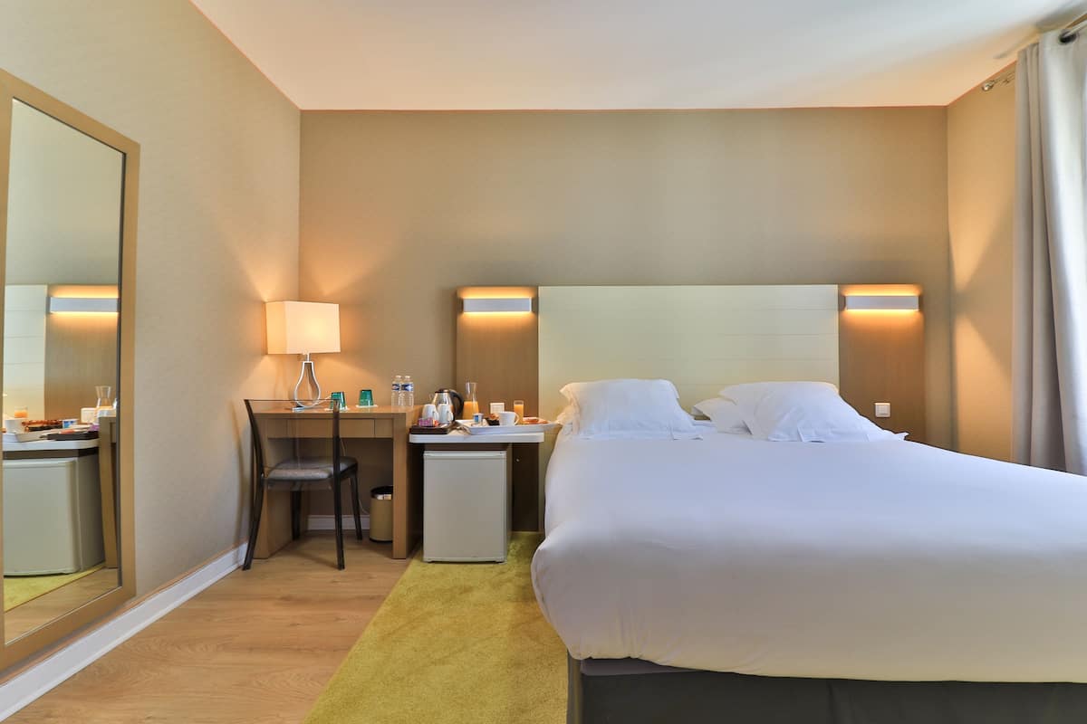 A hotel room with a bed, a desk, a chair, a small refrigerator, and a mirror.