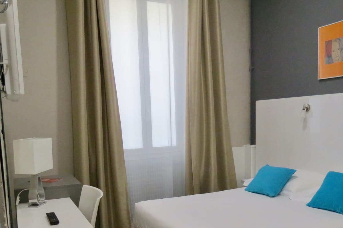 A hotel room with a bed, a desk with a lamp, a chair, and a window with curtains.
